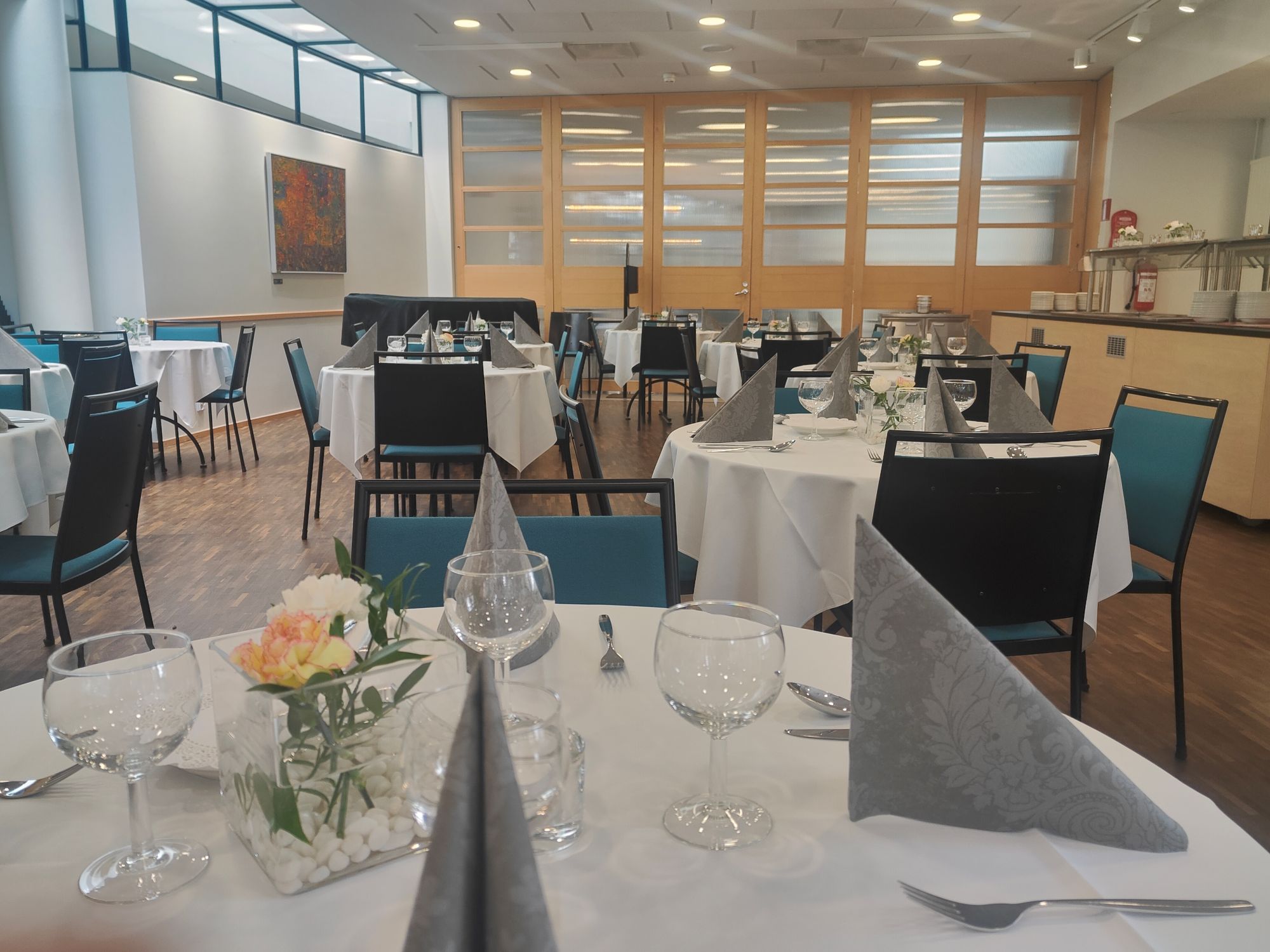 Metsäsali pöydillä valkoiset pöytäliinat, viinilasit, aterimet ja lautasliinat sekä pienet neilikat pöytäkukkina